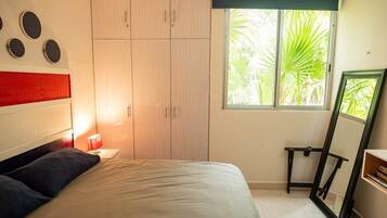 A bedroom with a bed, a wardrobe, a window with a view of palm trees, and a mirror.