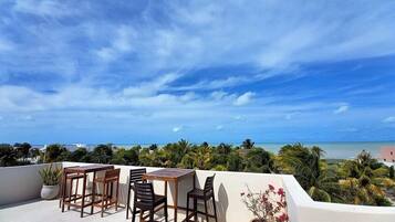 A rooftop terrace with a table and chairs, overlooking a beach and palm trees.