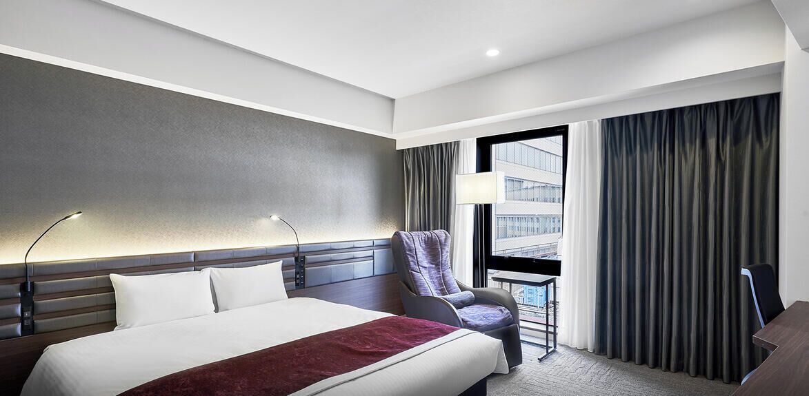 A hotel room with a bed, a chair, a desk, a window with curtains, and a view of a building.