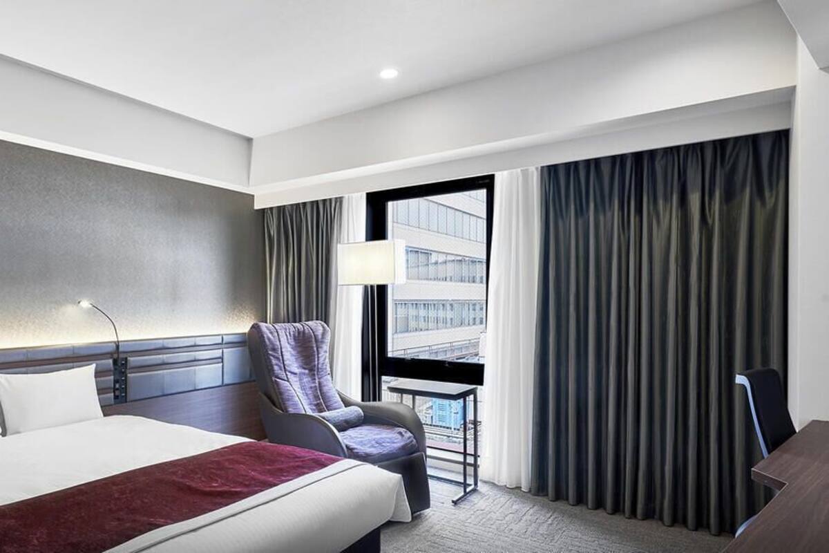 Habitación de hotel con cama, una silla, escritorio, ventana con cortinas y vista a un edificio.