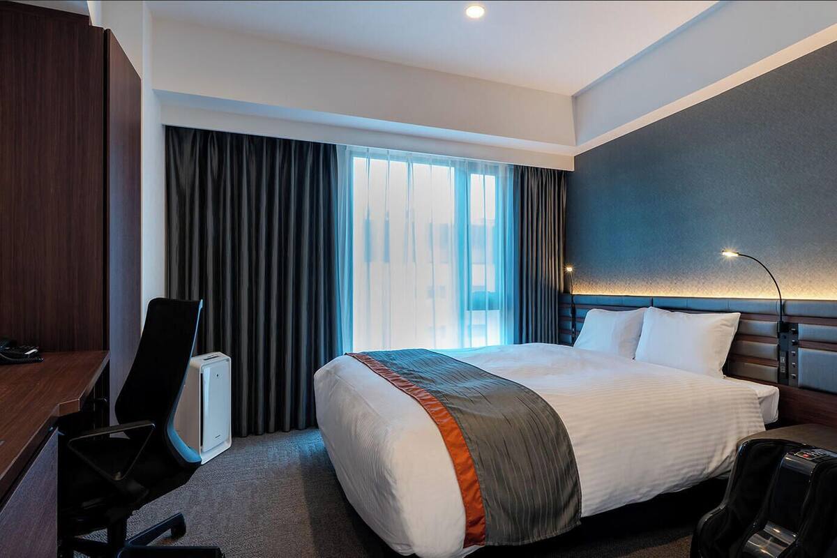 Una habitación de hotel con cama, escritorio, silla y un ventanal con cortinas.