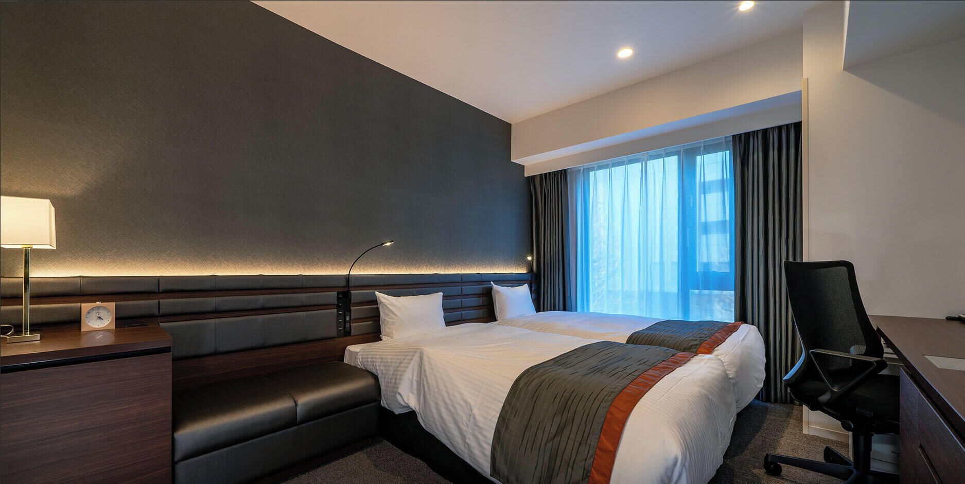 A modern hotel room with a large bed, a desk, a chair, and a window with curtains.