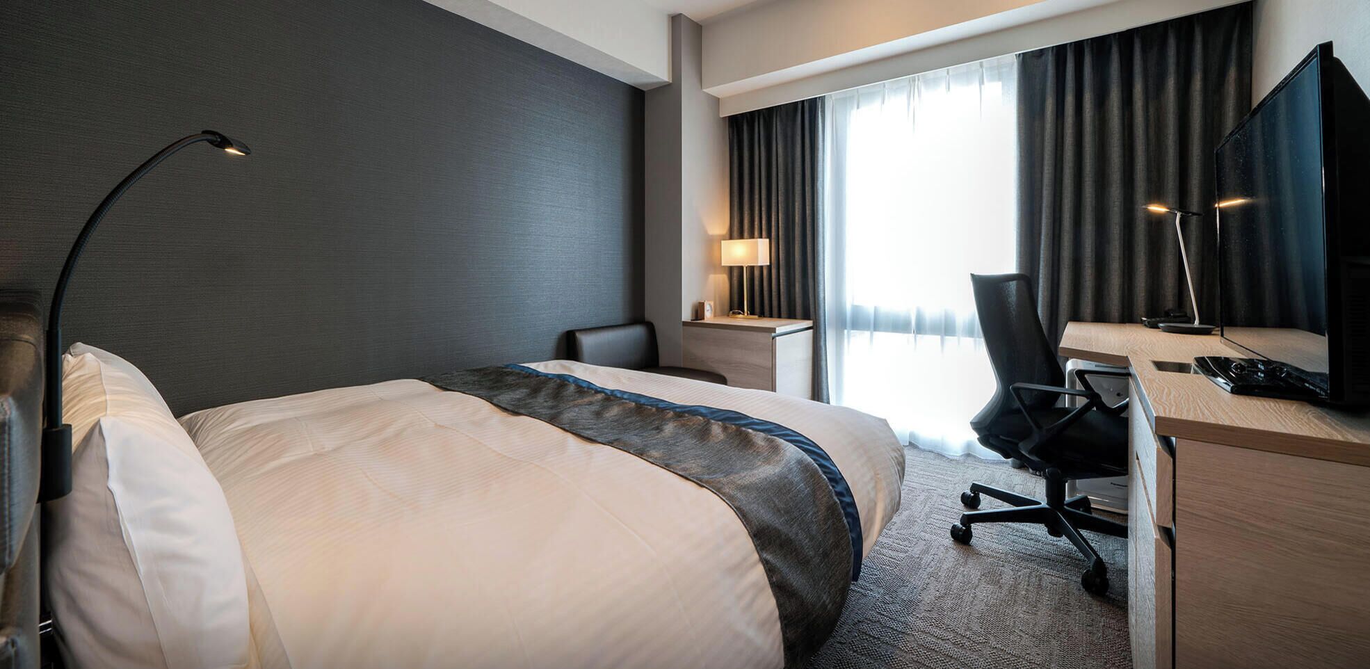 A hotel room with a bed, desk, chair, lamp, and a large window with curtains.
