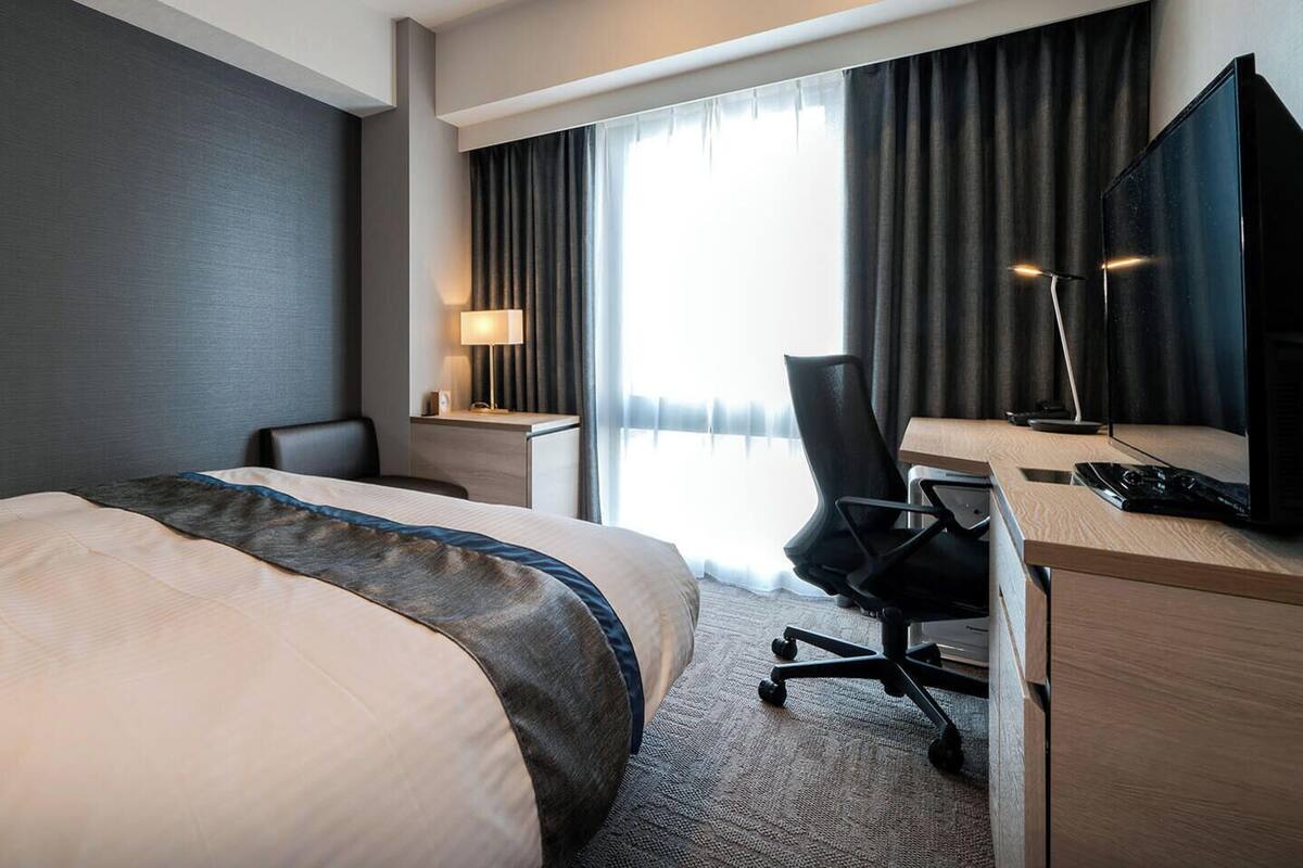 Una habitación de hotel con cama, escritorio, silla, lámpara y un ventanal con cortinas.