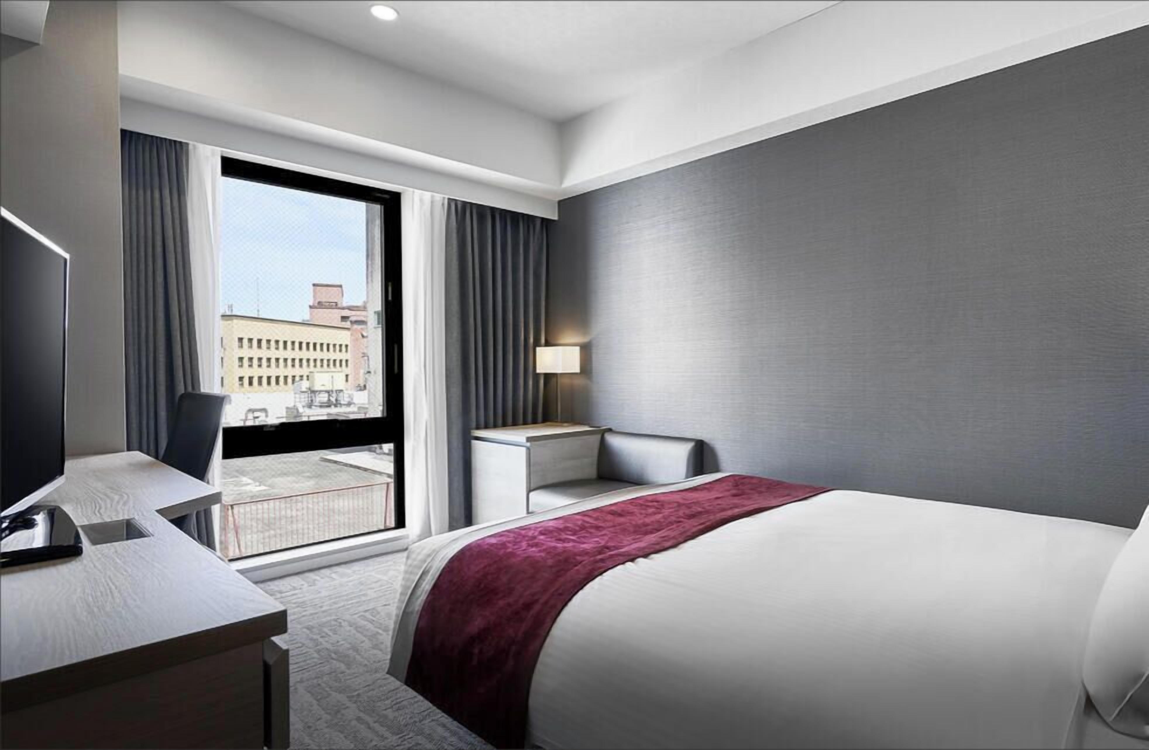 A modern hotel room with a large bed, a desk, a chair, and a view of the city through a large window.