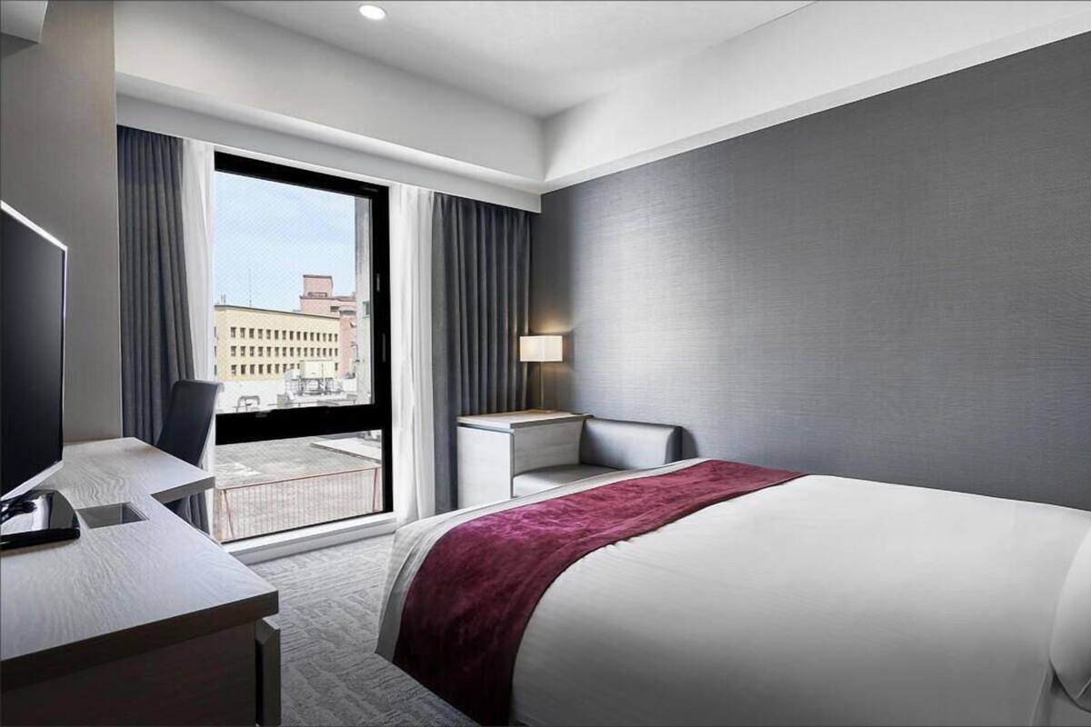 Habitación de hotel moderna con una cama grande, un escritorio, una silla y vistas de la ciudad a través de un amplio ventanal.