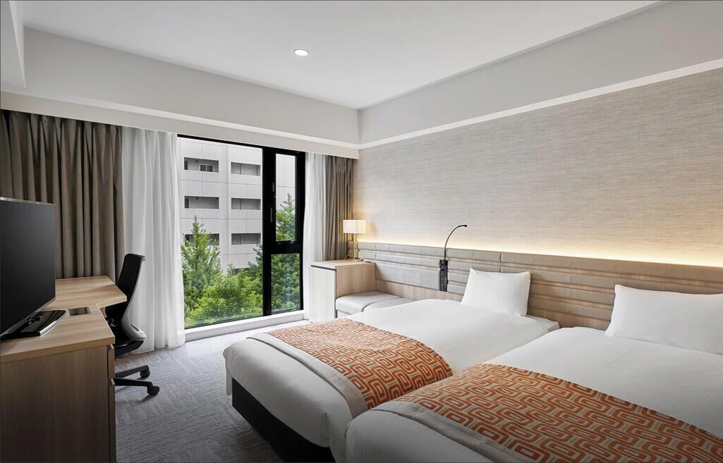 A hotel room with two beds, a desk with a TV, a lamp, and a view of greenery outside.