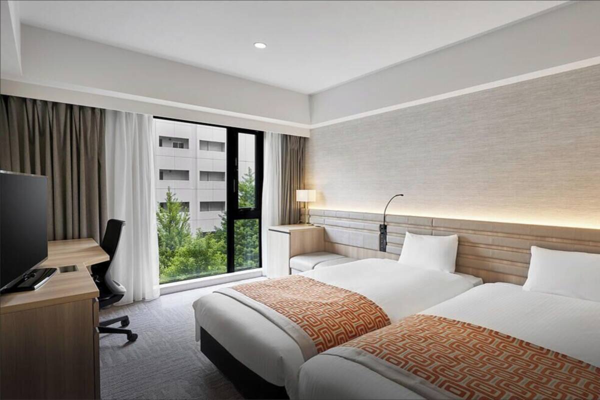Habitación de hotel con dos camas, un escritorio con televisor, una lámpara y vistas a zonas verdes.