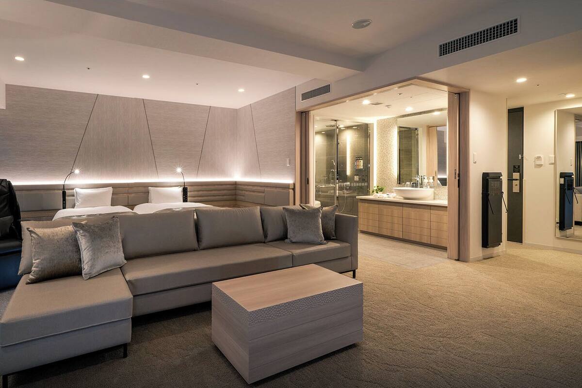 Habitación de hotel moderna con un amplio sofá en forma de L, un sillón independiente, una mesita, un vestidor, un baño con un espejo grande y zona de ducha.