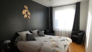Quarto com cama, duas cadeiras, mesa de cabeceira, luminária, objeto decorativo na parede e janela com cortinas.