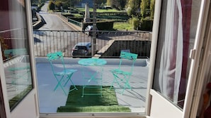 Quarto com varanda, cama com colcha florida e vista para a rua e vegetação lá fora.