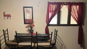 A dining area with a wooden table and four chairs, a framed picture on the wall, and a window with red curtains.