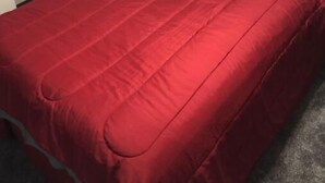 A bedroom with a red bedspread, a checkered pillow, a nightstand with a lamp, and a wall decoration of a bear.