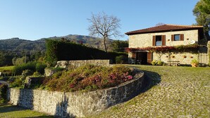 A stone house with a tiled roof, a garden with flowering plants, and a view of hills in the background.