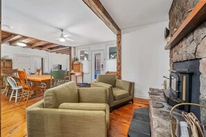 A cozy living room with a stone fireplace, wooden beams, and a dining area with a wooden table and chairs. - The Marigold PEC Family Farmhouse country escape (Prince Edward)