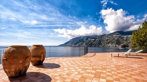 A terrace with tiled flooring, two large vases, and a railing overlooking a coastal view with mountains and a clear sky.
