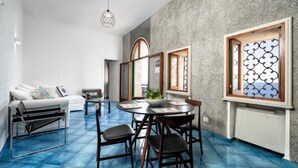 A modern living room with a blue-tiled floor, a white sofa, a dining table with chairs, and a kitchen area with a stove.
