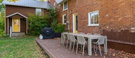 A backyard with a wooden deck, a barbecue grill, and a dining table with chairs.
