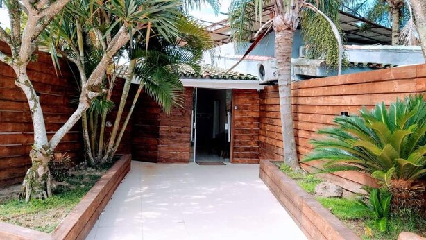 A wooden gate leading to a covered entrance, surrounded by palm trees and a small garden area. - Tropical Pousada (Guarujá)