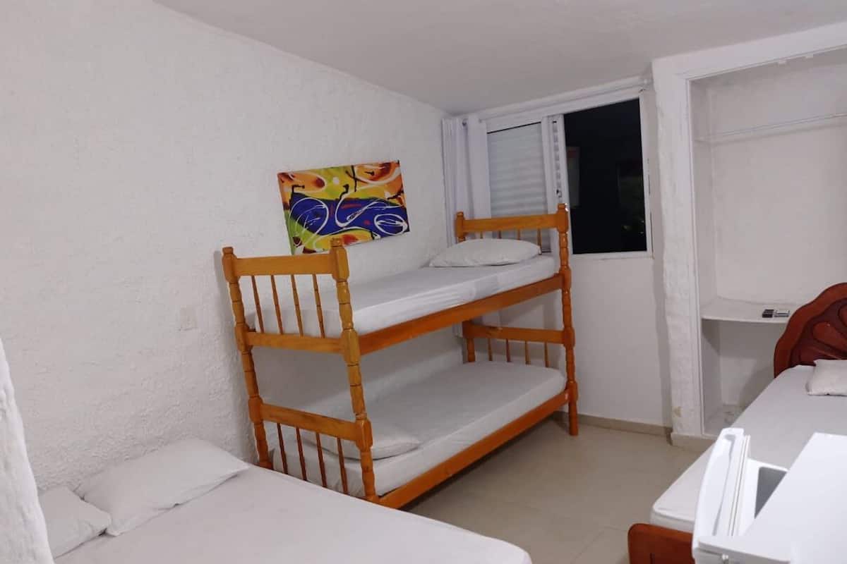 Habitación con literas, paredes blancas, una ventana con persianas, un cuadro de colores y una pequeña mesa con una silla.