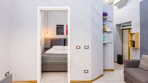 A hotel room with a bed, a sofa, a small table, a wall-mounted shelf with decorative items, and a door leading to another room. - Baubo - S. G. Armeno by Napoliapartments (Naples)