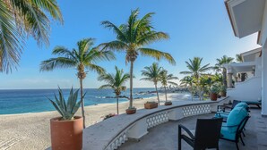 A beachfront patio with a view of the ocean, palm trees, and a seating area with chairs and a table. - Beachfront 5 BR Villas del Mar 461 fts. Private Pool, Hot Tub, WiFi + Much More! (Los Cabos)