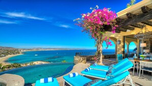 Zona junto a la piscina con sillones de descanso, vista al mar y un edificio con arcos.