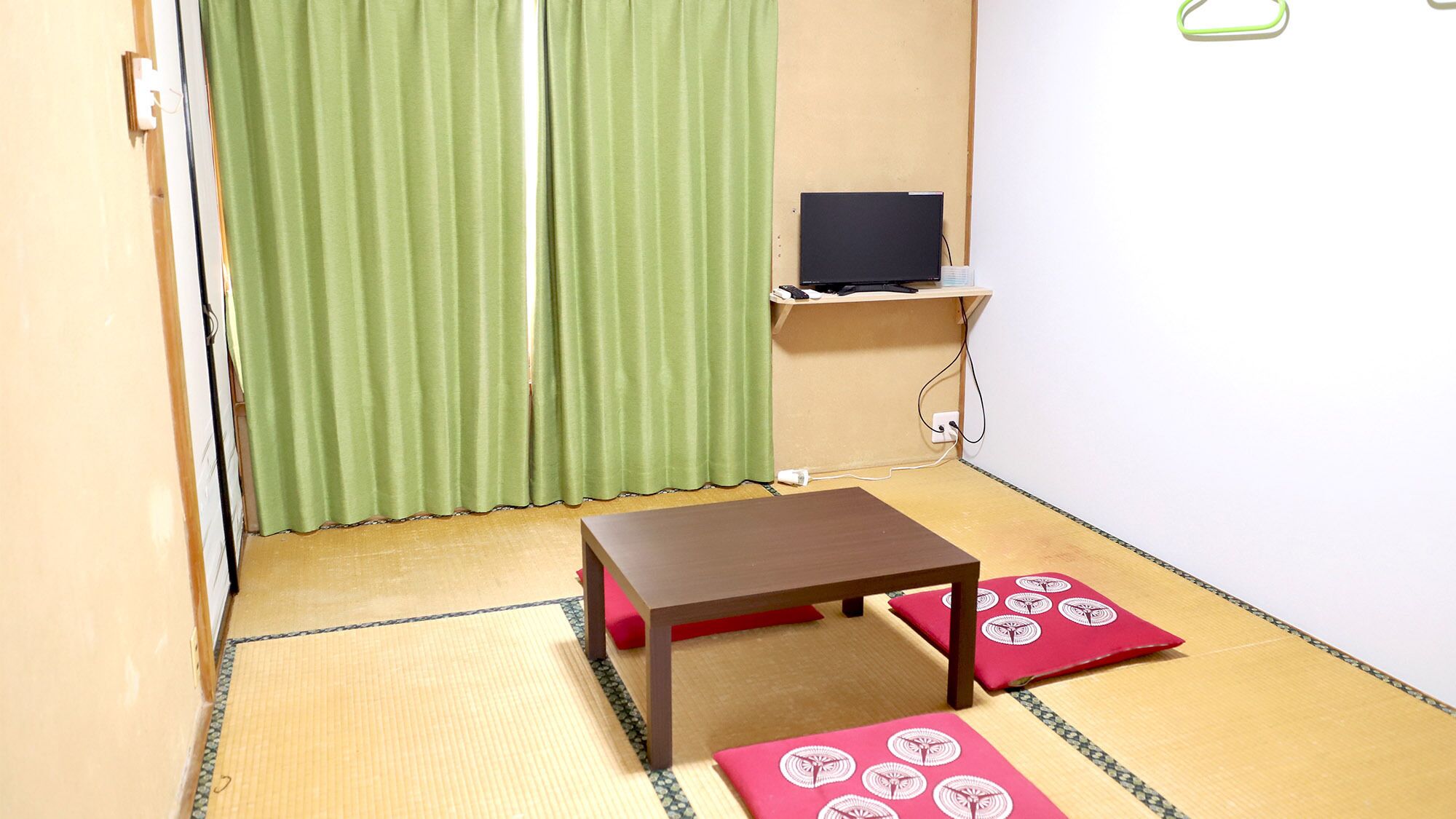 Una habitación con suelo de tatami, una pequeña mesa, un televisor en un estante y una cortina verde.