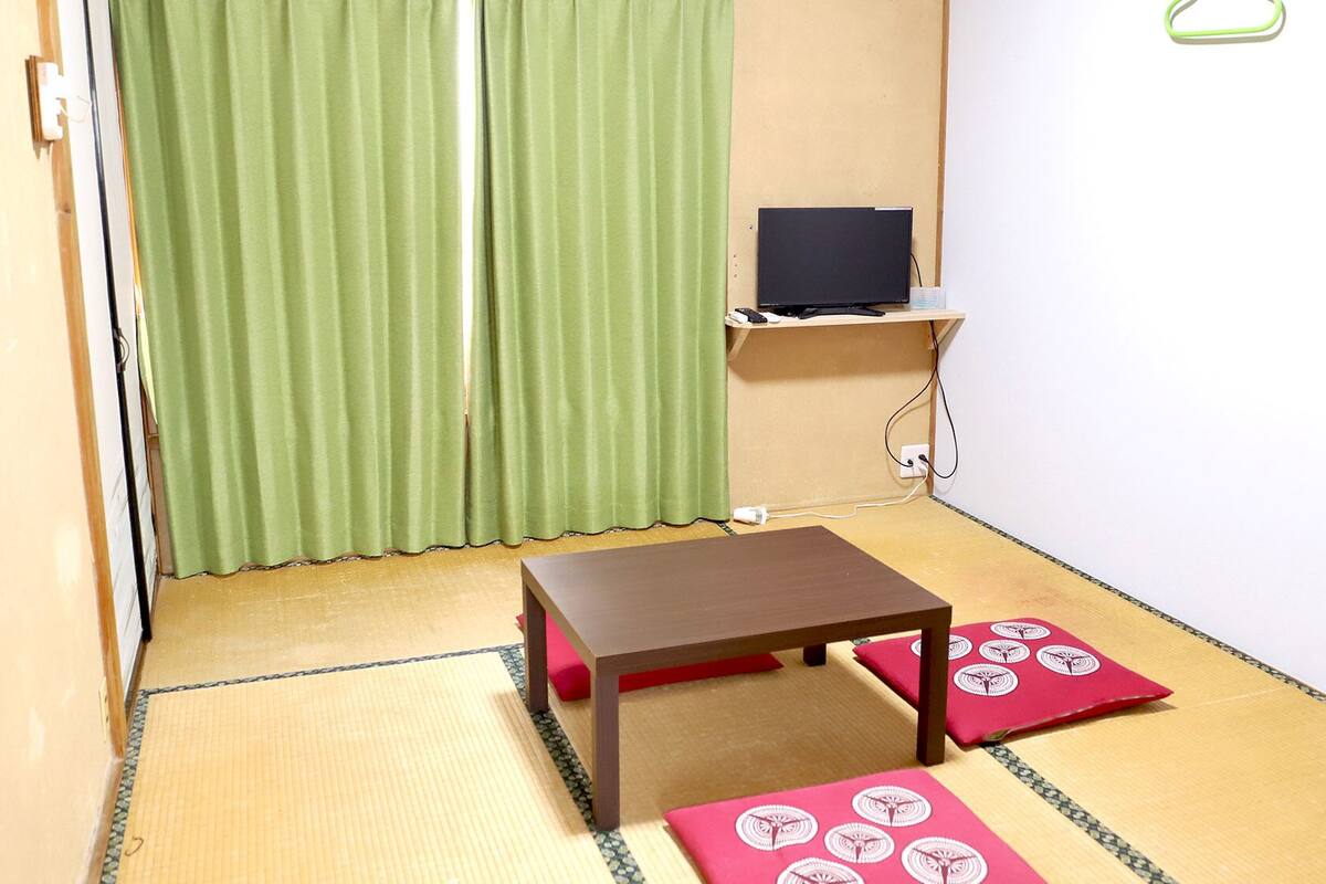 Una habitación con suelo de tatami, una pequeña mesa, un televisor en un estante y una cortina verde.