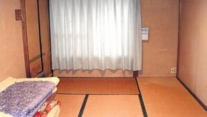 Una habitación con suelo de tatami, una cama con manta estampada, una cortina blanca y un equipo de aire acondicionado.