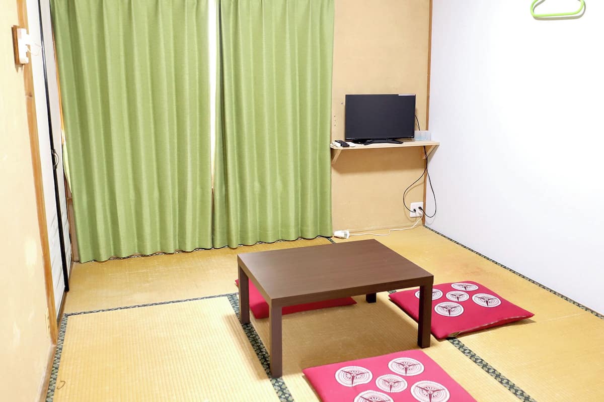 Una habitación con suelo de tatami, una pequeña mesa, un televisor en una estantería y una cortina verde.