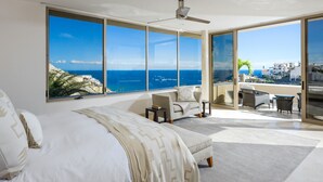 Un'ampia camera da letto con un letto grande, un'area salotto con divano e poltrone, e un balcone con vista sull'oceano e sugli edifici.