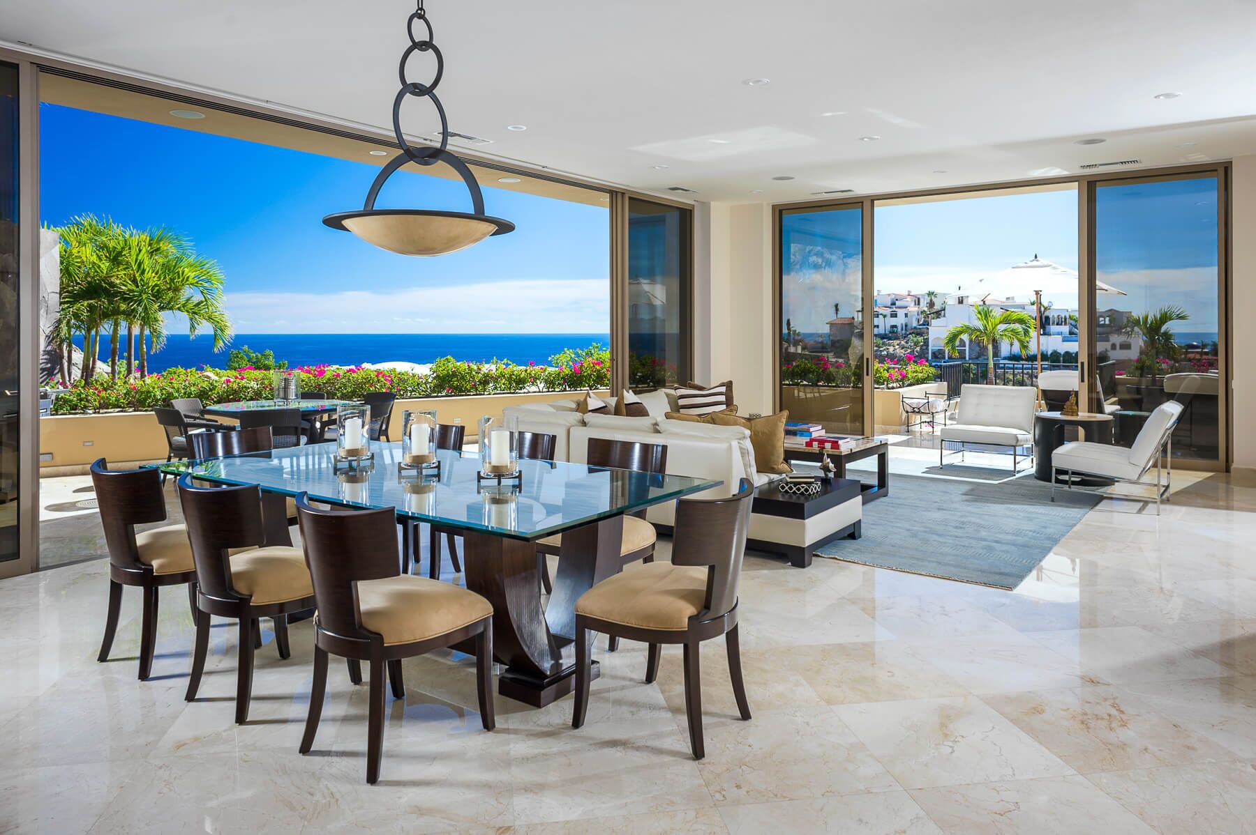 A spacious dining area with a glass table and chairs, a hanging light fixture, and a view of the ocean through large windows.