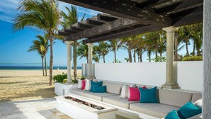 A beachside lounge with a white sofa set, teal and pink cushions, and a stone fire pit. - 2 FREE NIGHTS IN CABO at Villa Pacifica! (Cabo San Lucas)