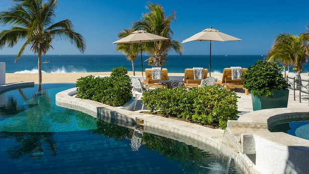 A poolside area with lounge chairs and umbrellas, a palm tree, and a view of the ocean. - 2 FREE NIGHTS IN CABO at Villa Pacifica! (Cabo San Lucas)