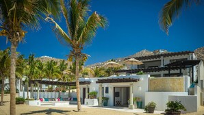 A resort with a sandy beach, palm trees, and a white building with a covered patio area. - 2 FREE NIGHTS IN CABO at Villa Pacifica! (Cabo San Lucas)