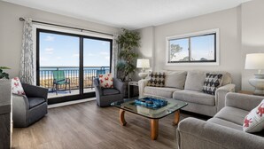 Una sala de estar con una mesa de centro de vidrio, dos sillones, un sofá y vistas a la playa a través de amplias ventanas.