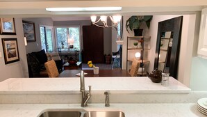 A modern kitchen with a double sink, marble countertops, and a view into a living room with a sofa and a dining area.