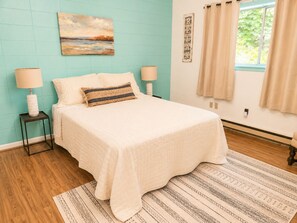 Quarto com cama, duas luminárias de cabeceira, um quadro na parede, janela com cortinas e um tapete estampado.