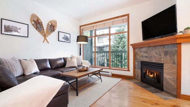 A modern living room with a leather sofa, a coffee table, a fireplace, and a large window with a view of trees.