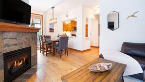 A modern living room with a fireplace, a dining area, and a kitchen visible through an open doorway.
