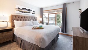 A modern hotel room with a large bed, a flat-screen TV, a bedside table with a lamp, and a window with curtains.