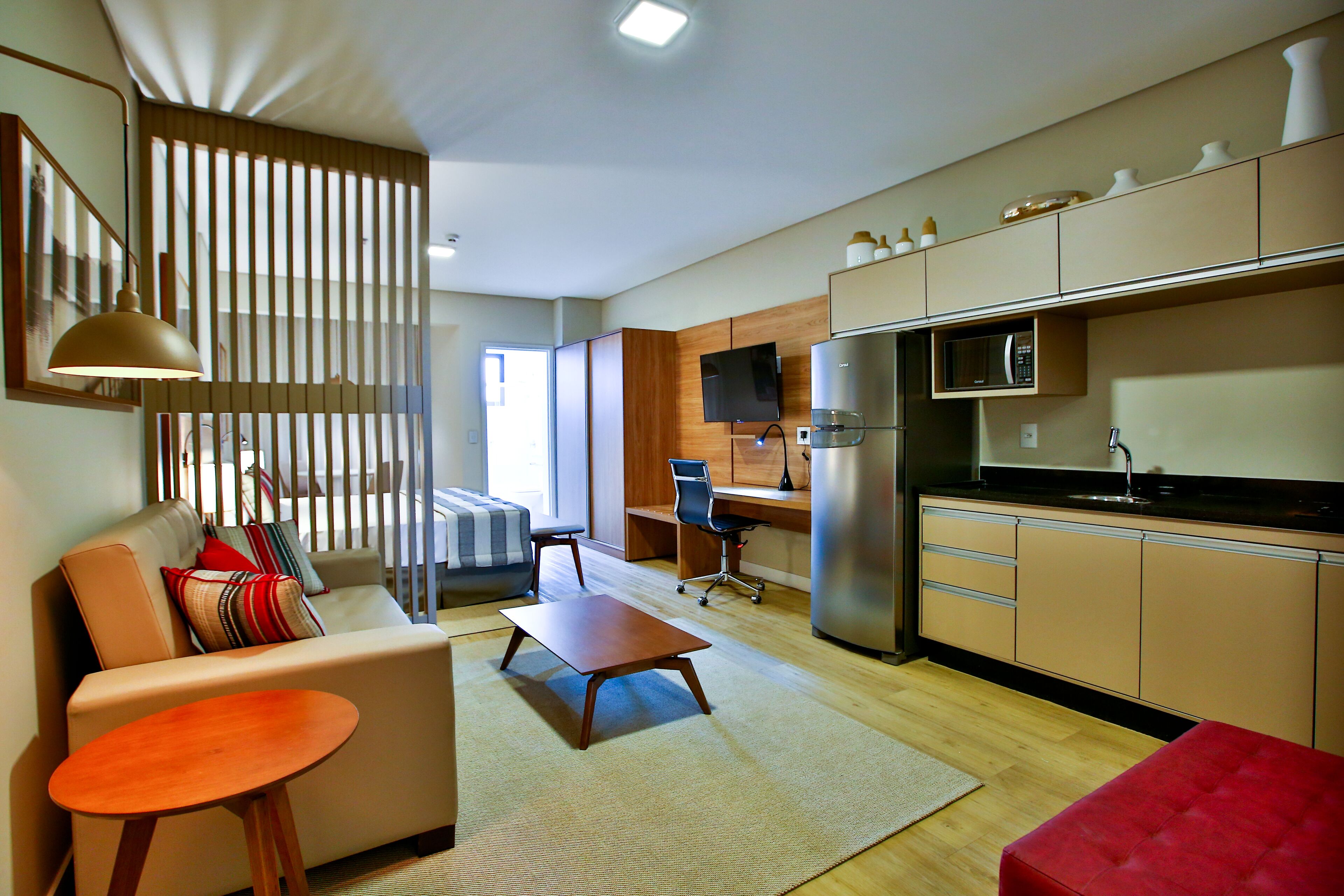 A modern living room with a sofa, coffee table, and a red ottoman. A kitchen with a microwave and refrigerator is visible in the background.
