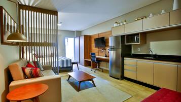 A modern living room with a sofa, coffee table, and a red ottoman. A kitchen with a microwave and refrigerator is visible in the background.