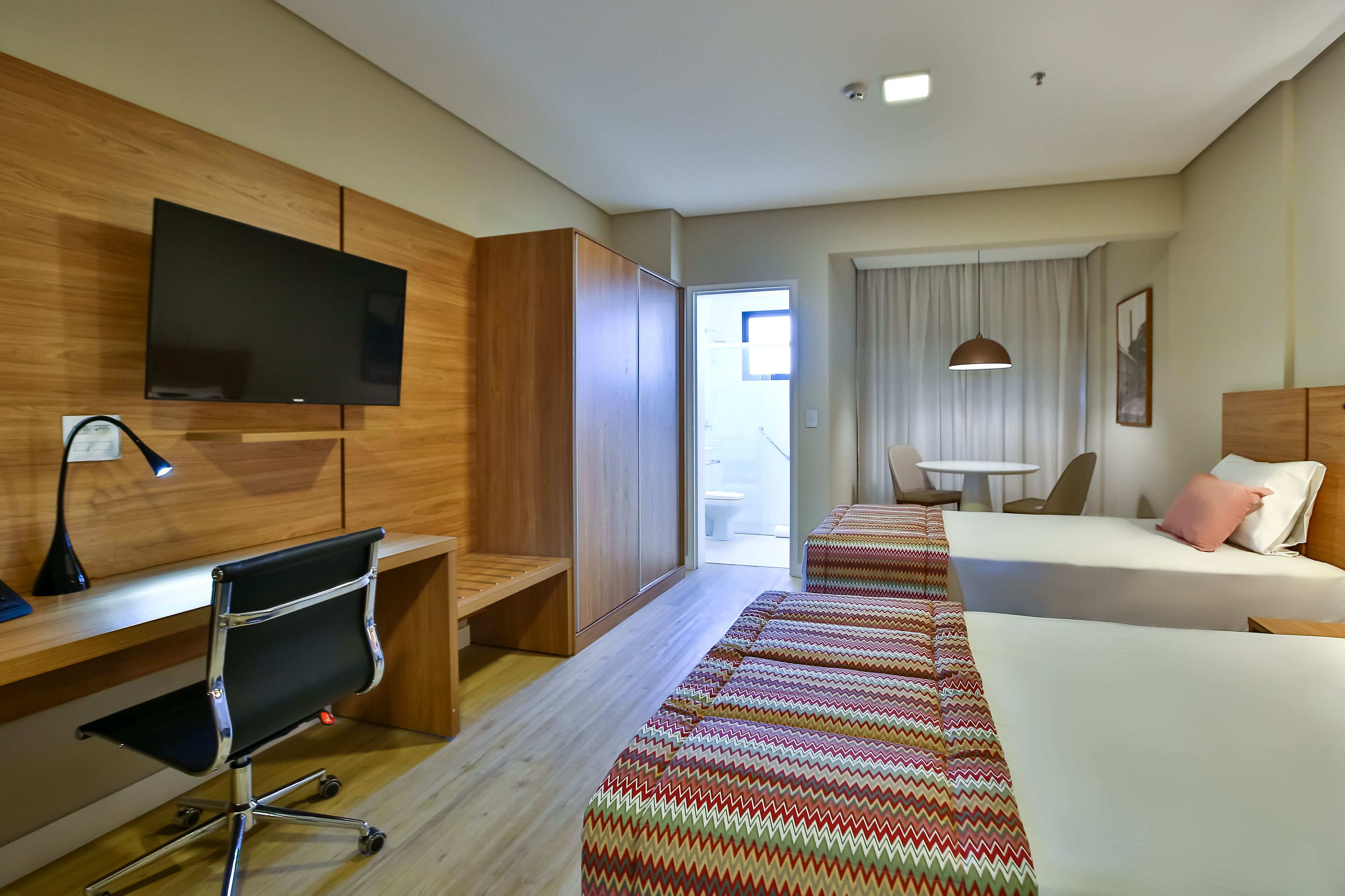 A hotel room with a wooden desk, a chair, a TV, a bed with a colorful blanket, a bedside table, and a window with curtains.