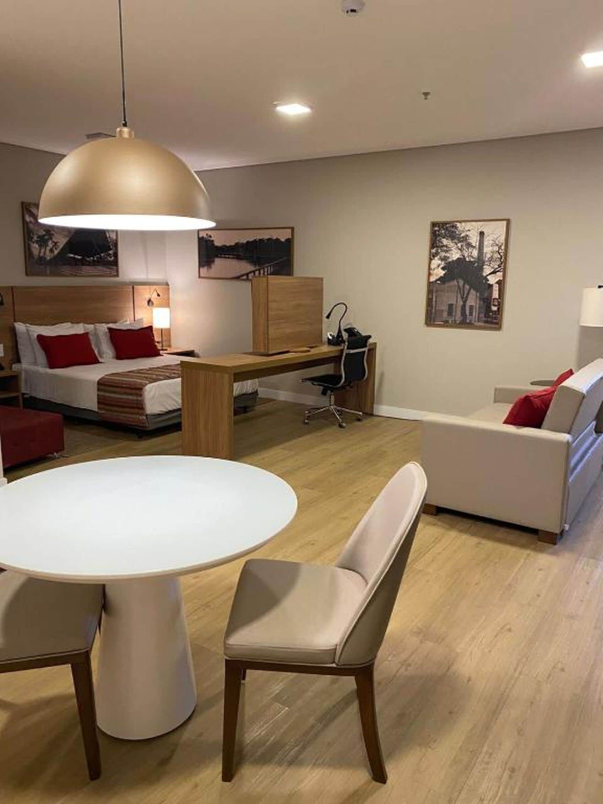 A modern living room with a round dining table, a sofa, a desk, and a chair. There are two paintings on the wall and a ceiling light.
