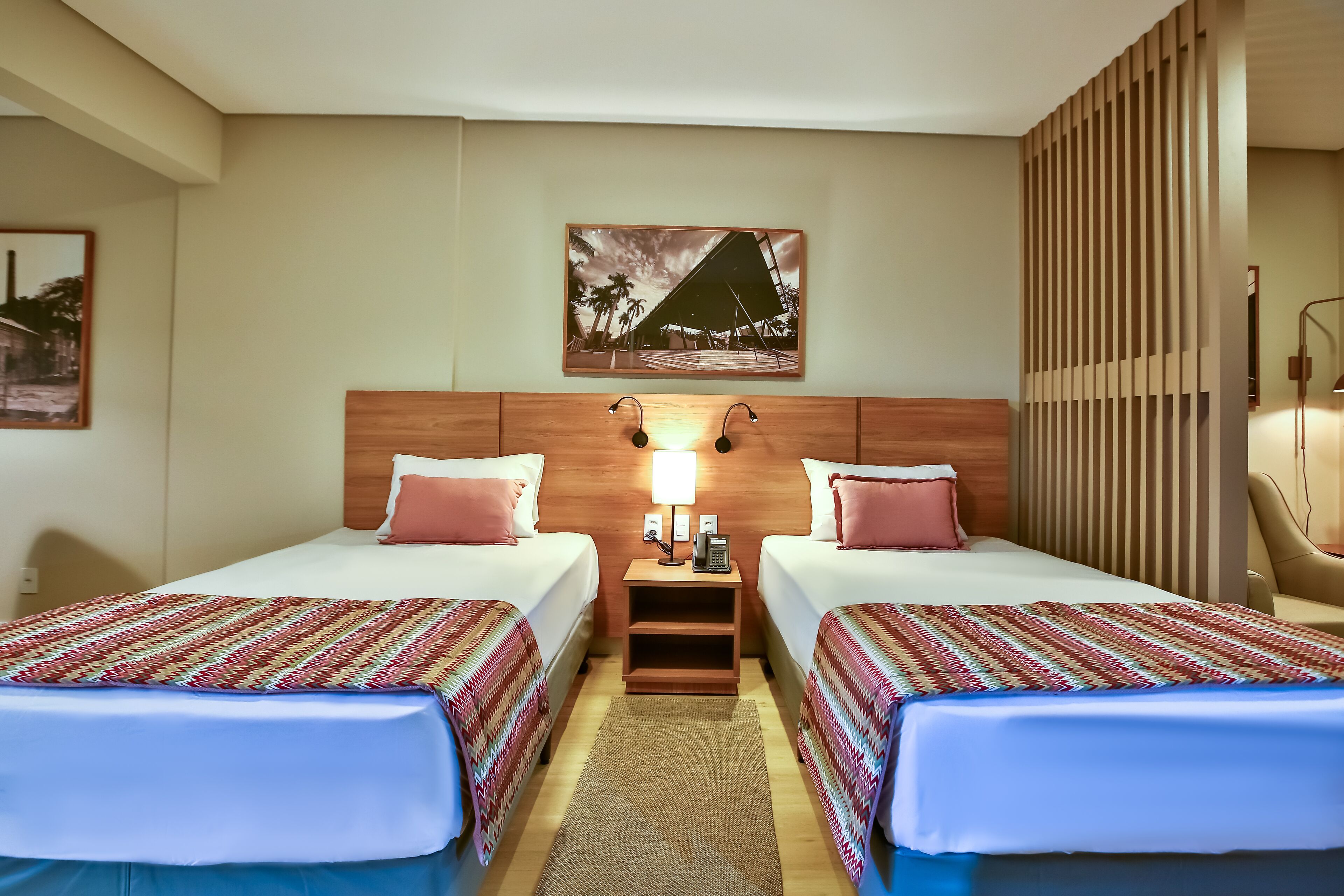 A hotel room with two beds, a nightstand with a lamp, a painting on the wall, and a wooden partition.