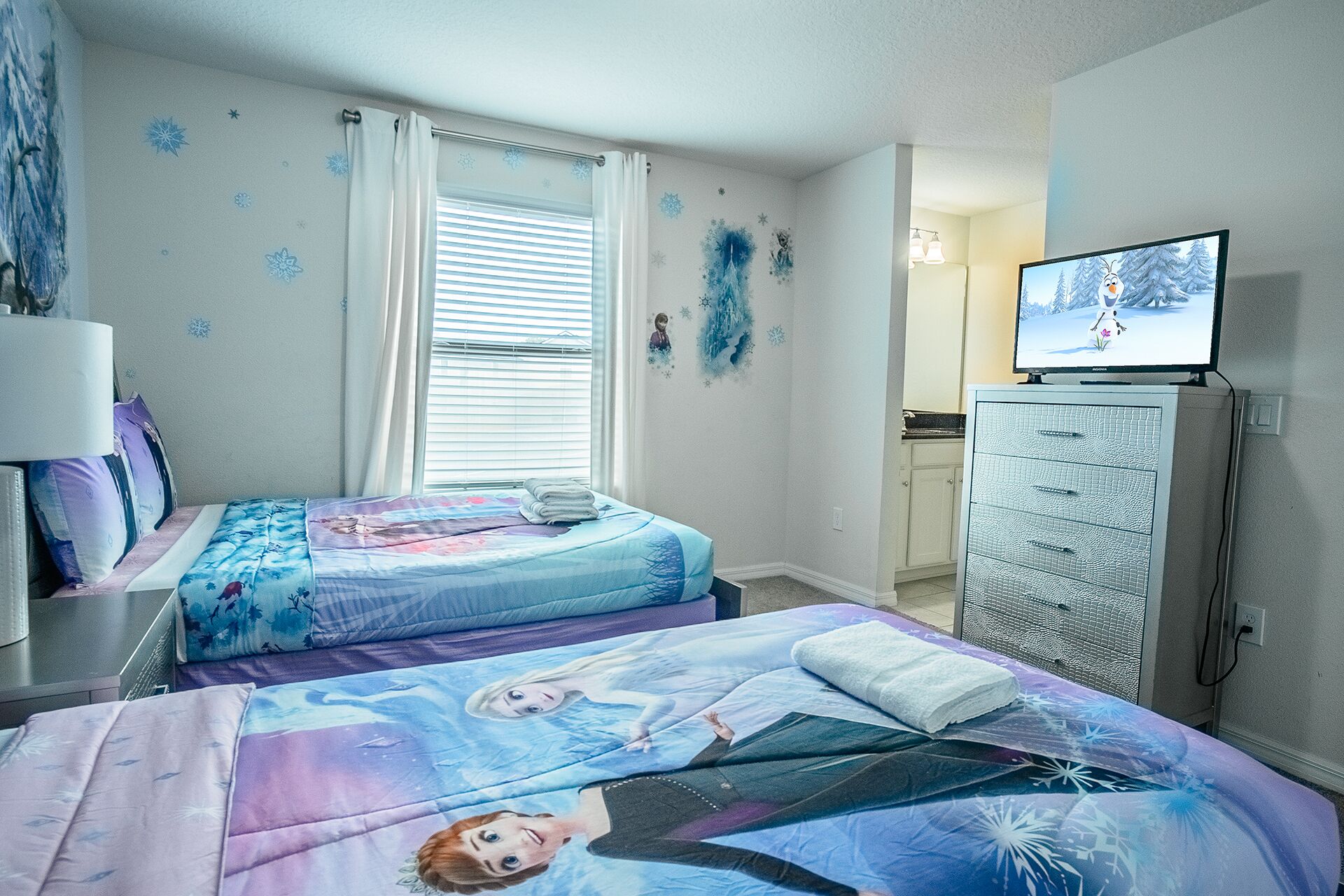 A bedroom with two beds, a dresser, a television, and a window with blinds.
