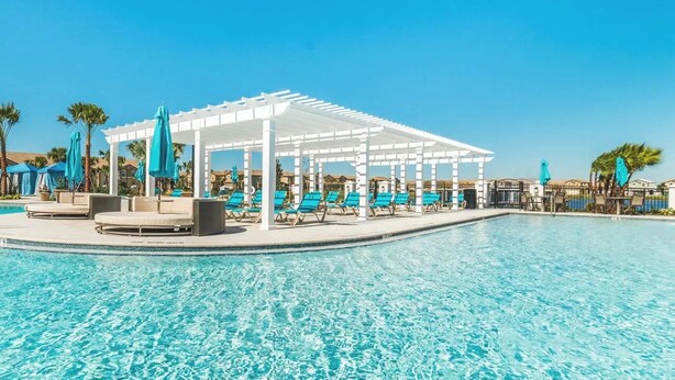 Uma área de piscina com pergolado branco, cadeiras de lounge e palmeiras.