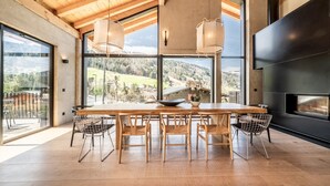 A modern dining area with a wooden table and chairs, large windows offering a view of the outdoors, and a built-in fireplace. - NELCOTE - Design and luxury chalet with hot tub (Morzine)